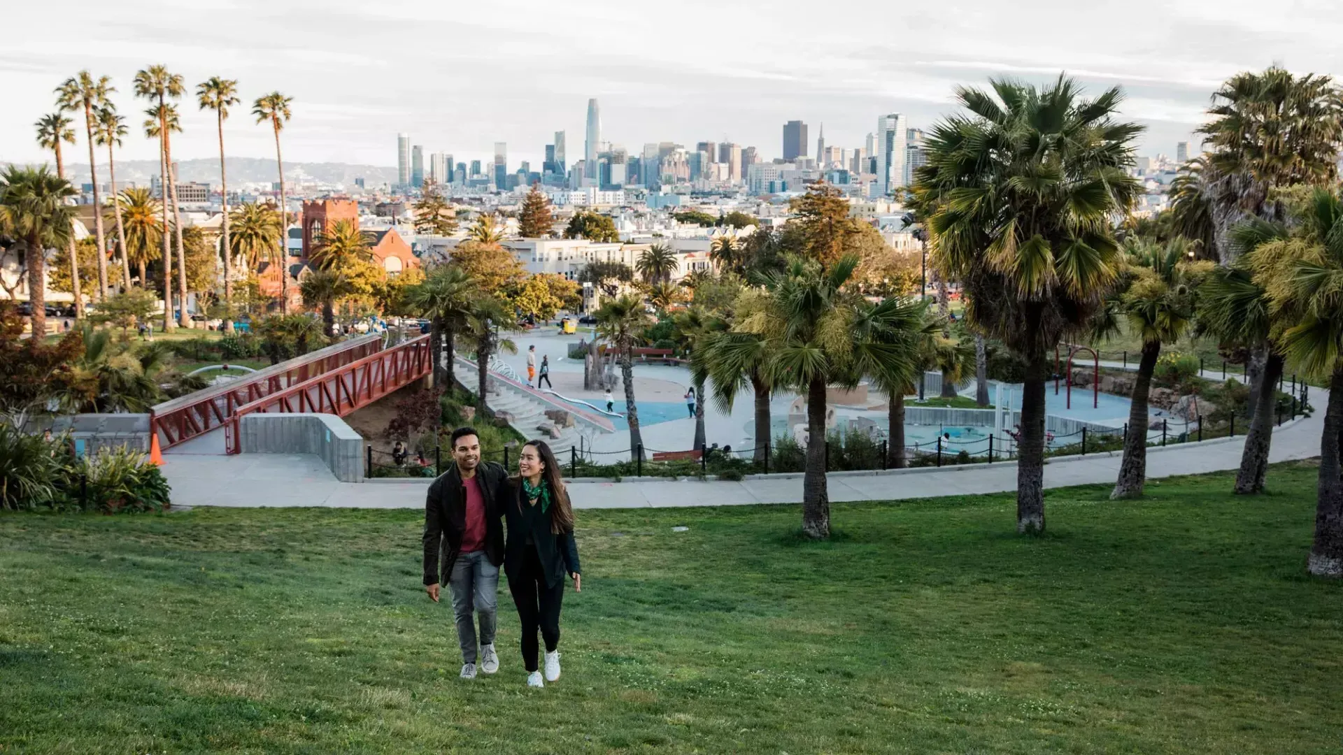 Ein Paar geht auf die Kamera zu, hinter ihnen Dolores Park und die Skyline von San Francisco.